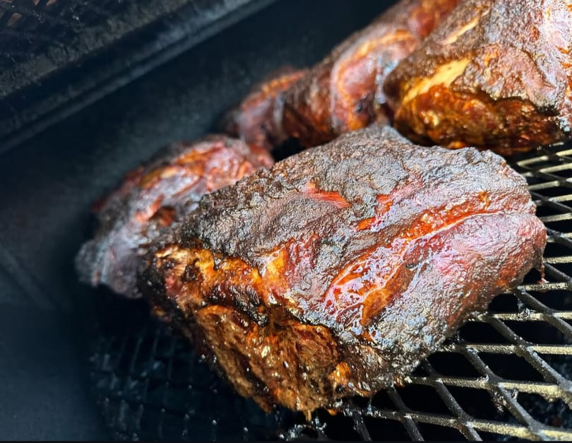 Close-up of several large cuts of barbecued meat with a dark, crispy crust on a grill.