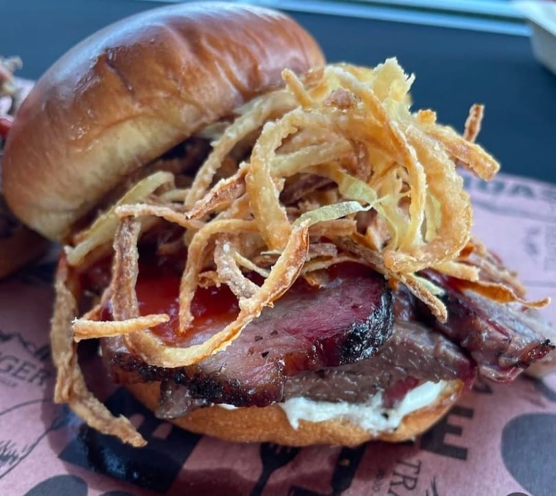 Close-up of a brisket sandwich with crispy fried onions, barbecue sauce, and white sauce.